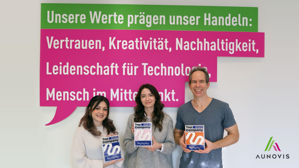Gruppe von drei Personen hält Auszeichnungen für Top Company 2023, 2024 und 2025 vor einer Wand mit einer inspirierenden Botschaft, die die Werte Vertrauen, Kreativität, Nachhaltigkeit und Leidenschaft für Technologie betont.